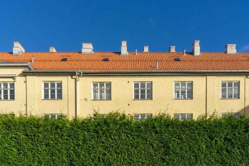 En gul leiegård med rødt teglsteinstak bak en tett grønn hekk
