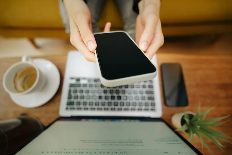 To hender holder en mobiltelefon over en åpen laptop på et bord, med en kaffekopp ved siden av