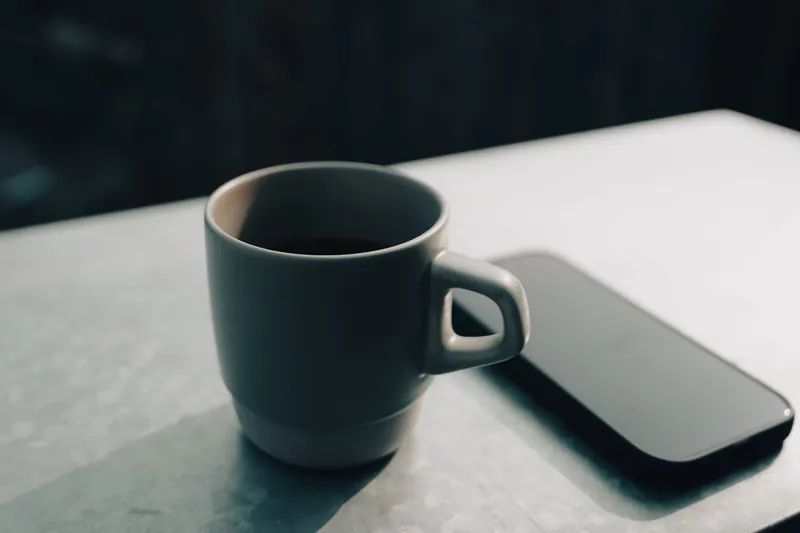 En grå keramikkopp med kaffe ved siden av en mobiltelefon med mørk skjerm på et lyst bord