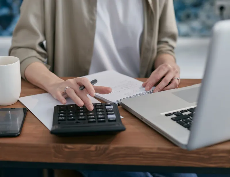 En person regner på en kalkulator ved et skrivebord med laptop, notatblokk og kaffekopp