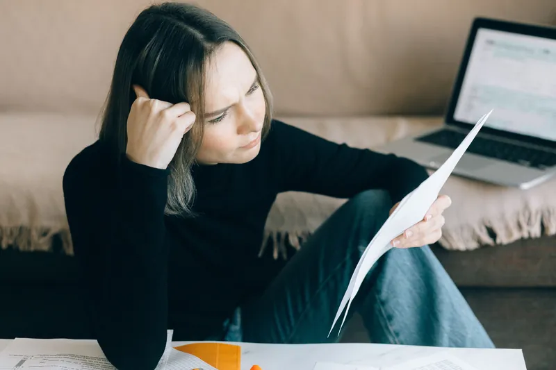 En kvinne sitter på gulvet ved sofaen og ser bekymret på et ark, med en laptop bak seg