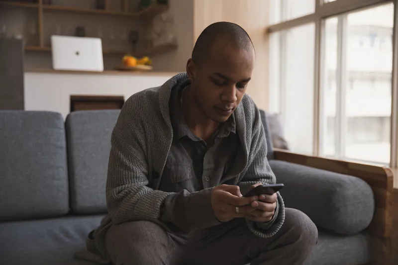 En mann sitter i en grå sofa og leser på mobilen sin, med et vindu i bakgrunnen