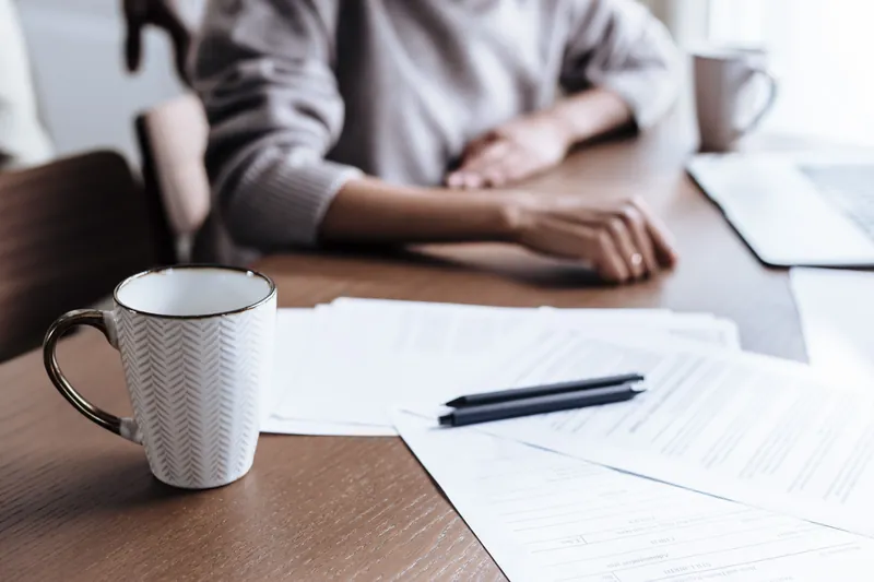 Papirer, penn og en kaffekopp på et spisebord, med en person utenfor fokus i bakgrunnen