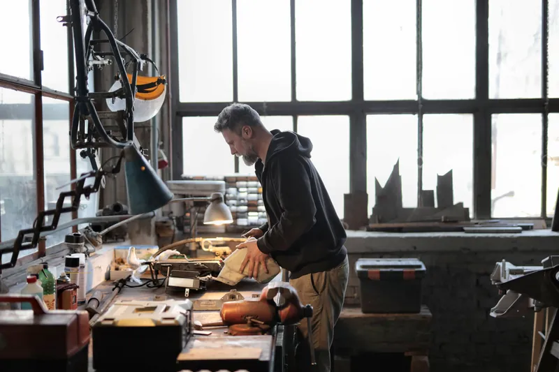 En mann står alene ved arbeidsbenken i sitt eget verksted og arbeider med en metalldel
